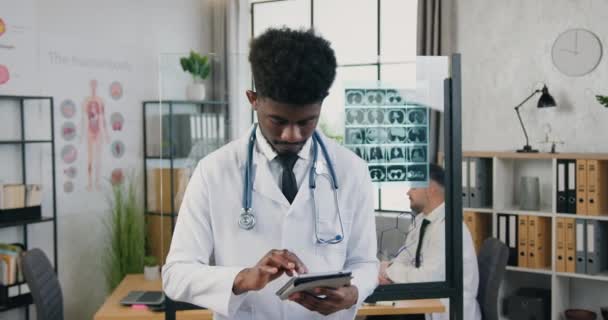 Porträt eines gut aussehenden, fröhlichen jungen, qualifizierten, schwarzhäutigen Arzthelfers, der sein Tablet-Gerät benutzt und in die Kamera in der modernen Klinik blickt — Stockvideo