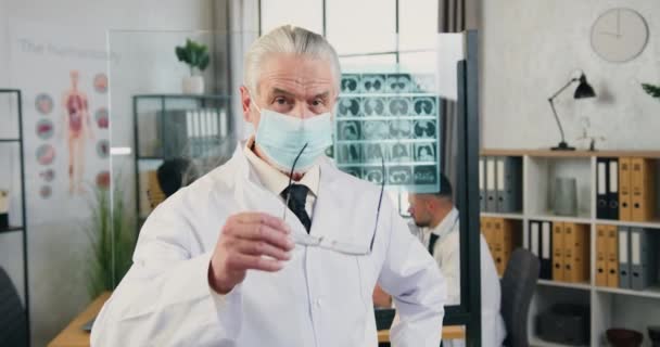 Detailní záběr sebevědomého uznávaného zralého šedovlasého lékaře v masce obličeje, který si nasazuje brýle pózující na kameru v moderní lékařské ordinaci, covid-19 pandemie koncept — Stock video