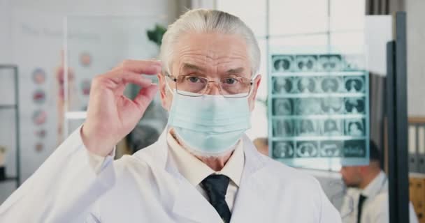 Retrato de respetado médico masculino inteligente de pelo gris con máscara médica que se quita las gafas posando en cámara en el consultorio médico moderno — Vídeos de Stock