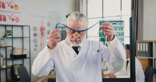 Atraktivní sebevědomý uznávaný kvalifikovaný starší vousatý doktor si nasazuje stetoskop kolem krku a dívá se do kamery v moderní nemocniční kanceláři, pohled zepředu — Stock video