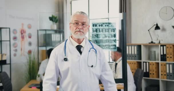 Portrait de beau sérieux expérimenté respecté médecin barbu de 65 ans dans des lunettes en manteau blanc avec stéthoscope qui pose sur la caméra avec les bras croisés dans la salle de travail de l'hôpital — Video