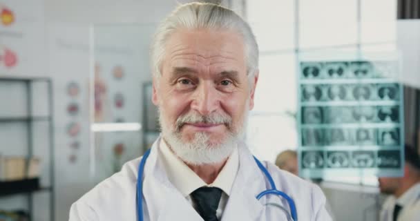 Detailní portrét dobře vypadajícího pozitivního zkušeného šedovlasého vousatého doktora, který se dívá do kamery s upřímným úsměvem v nemocniční dílně — Stock video
