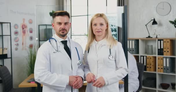 Atraente confiante experiente bem sucedido médicos do sexo masculino e feminino em uniformes hospitalares em pé na frente da câmera na moderna sala de trabalho clínica — Vídeo de Stock