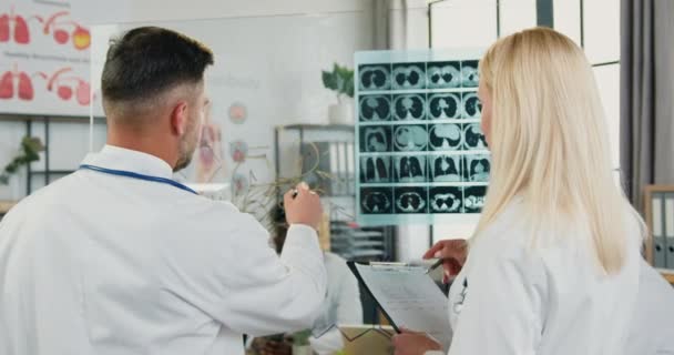 Medicinkoncept där snygga smarta kvalificerade vuxna manliga och kvinnliga läkare står nära glasvägg i arbetsrummet och diskuterar resultat av röntgenundersökning och ritning kemisk formel på denna vägg — Stockvideo