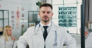Yakışıklı, başarılı, sakallı, üniformalı bir doktor. Kameranın önünde modern tıp laboratuarında meslektaşlarının geçmişinde çapraz kollarla duruyor.