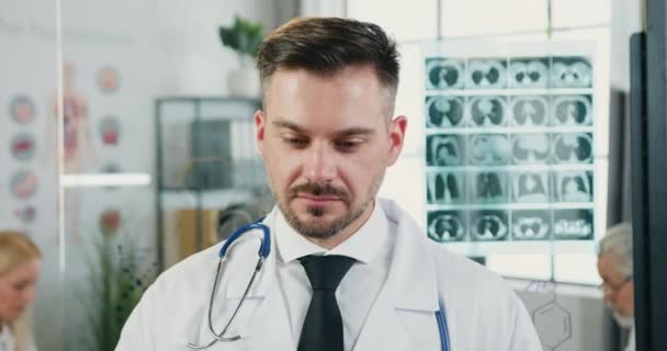 Nahaufnahme Porträt eines gut aussehenden lächelnden zufriedenen erwachsenen bärtigen Arztes, der im modernen Krankenhauszimmer eine Brille aufsetzt, wenn er in die Kamera schaut — Stockvideo