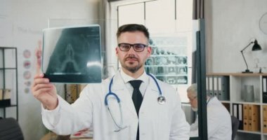 Güzel görünüşlü, deneyimli, sakallı, 40 yaşında, gözlüklü, klinikteki çalışma odasında röntgen taramasıyla çalışan kendine güvenen bir doktor.