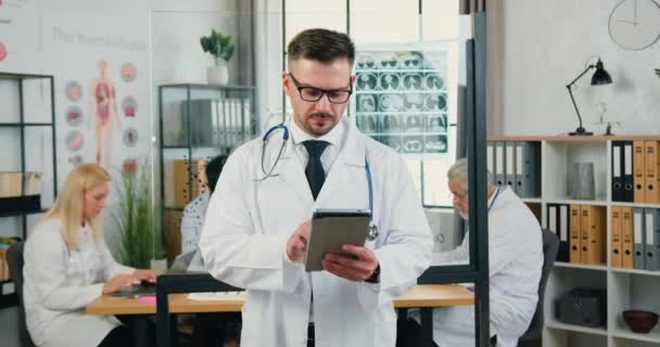 Portret sympatycznego doświadczonego dorosłego, brodatego lekarza w białym płaszczu, który korzystając z tabletu w gabinecie kliniki przy pracownikach omawiających momenty pracy — Wideo stockowe