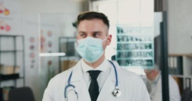 Kamera, modern hastane çalışma odasında kameraya poz veren, salgın hastalıklı covid-19 zamanında korunan koruyucu maskeli başarılı yetişkin doktorlara çevriliyor.