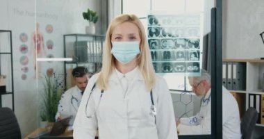 Kendine güveni yüksek, koruyucu maskeli güzel sarışın kadın doktorun portresi modern klinik çalışma odasındaki kameraya, çalışma akışına, tıbba ve covid-19 konseptine işaret ediyor.