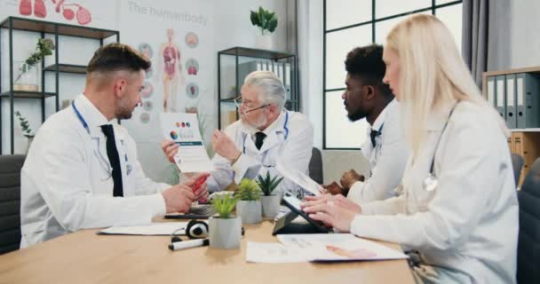 Bonito sério respeitado cabeça barbuda médico explicando resultados de pesquisas de DNA usando relatório com gráficos para seus confiantes experientes colegas multirraciais masculinos e femininos no consultório médico — Vídeo de Stock