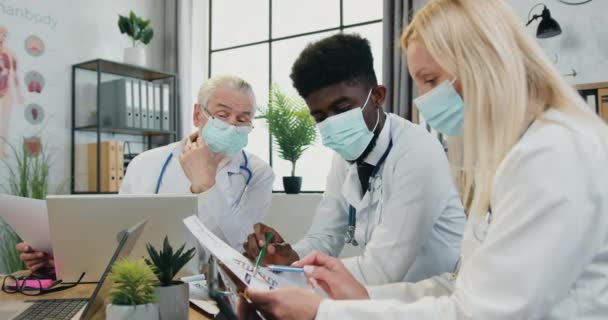 Medische bijeenkomst tijdens coronavirus pandemie waar aantrekkelijke slimme succesvolle bekwame mannelijke en vrouwelijke artsen in maskers bespreken medische momenten in hun werk tijdens overleg in kliniek kantoor — Stockvideo