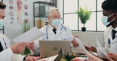 Kendine güveni tam başarılı yüz maskeli baş doktor modern klinik ofisindeki çok kültürlü meslektaşlarıyla birlikte danışmanlık yapıyor.