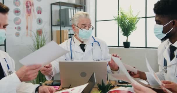 Good-looking confident successful respected head doctor in face mask holding consultation together with his skilled multiracial colleagues in modern clinic office,front view — Stock Video