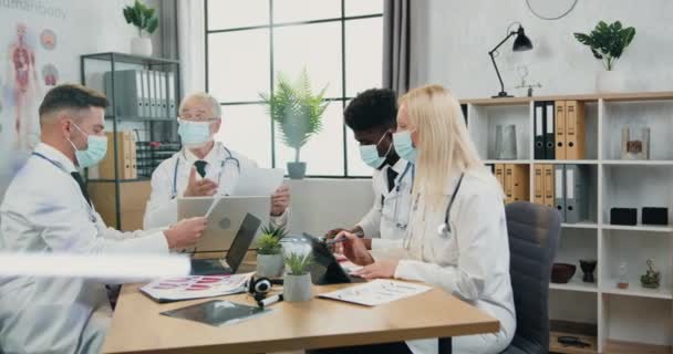 Flujo de trabajo en el hospital durante covid-19 donde atractivos médicos cualificados inteligentes y seguros en máscaras faciales trabajando juntos en la sala de juntas y discutiendo momentos médicos de trabajo — Vídeo de stock