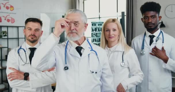 Szerethető pozitív képesített különböző életkorú férfi és női orvosok a kamera előtt állnak a modern orvosi szobában, elölnézetből — Stock videók
