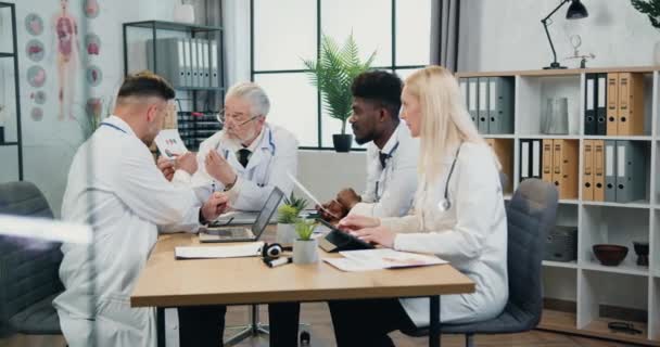 Приятные умные квалифицированные многорасовые врачи беседуют со своим уважаемым бородатым 65-летним главным врачом во время совместной встречи в медицинском кабинете, концепция медицины — стоковое видео
