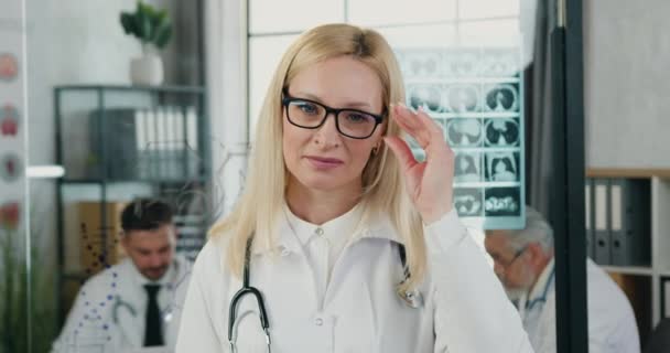 Charmigt leende högkvalificerad blond kvinnlig läkare tar av glasögon och tittar in i kameran med nöjda ansiktsuttryck i moderna medicinska kontor, medicin koncept — Stockvideo