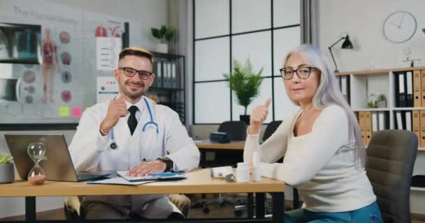 Aantrekkelijke glimlachende ervaren bebaarde arts en zijn behoorlijk volwassen vrouwelijke patiënt die duimen laat zien in camaera tijdens haar bezoek aan de kliniek — Stockvideo