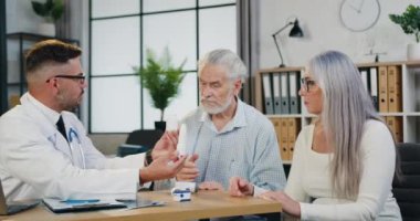 Yakışıklı, kendine güvenen, sakallı, gözlüklü bir doktorun yetişkin çiftlere tıbbi muayenehane ziyareti sırasında ilaç verdiği klinikte randevu.