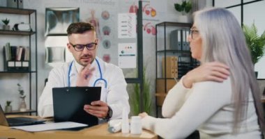 Çekici, zayıf, kır saçlı kadın özel ofisinde yetenekli sakallı doktorla konuşuyor ve sağlık şikayetlerini anlatıyor.