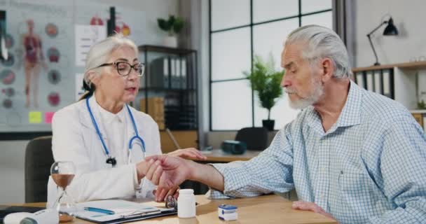Скорее всего, обеспокоенный старший бородатый мужчина посещает медицинский кабинет, где красивая заботливая квалифицированная женщина-врач проверяет его пульс, кладя пальцы на его запястье — стоковое видео