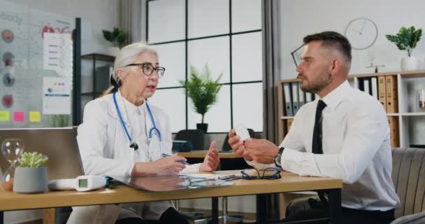 Fešák oslabený dospělý vousatý muž dostal pilulky od zkušené starostlivé starší doktorky a naslouchat doporučení, jak je vzít, pohled zepředu, zpomalení — Stock video
