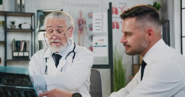 Pohledný pozitivní sebevědomý 40-ti letý vousatý muž na návštěvě lékařské ordinace, kde zodpovědný zkušený šedovlasý lékař diskutuje s ním výsledky rentgenového skenování — Stock video