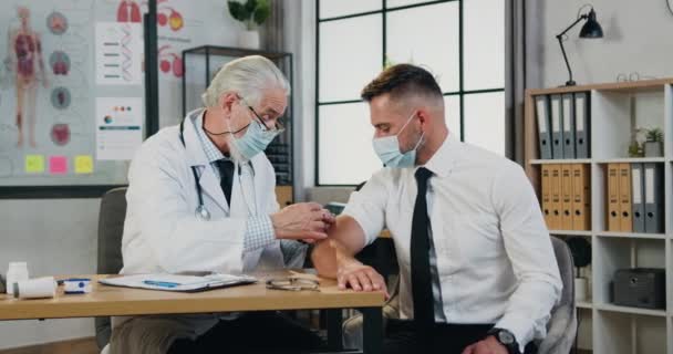Attrayant responsable senior cheveux gris médecin masculin dans le masque faire injection de vaccin de covide-19 à l'homme adulte confiant positif dans le masque médical dans le bureau de la clinique, la prévention à temps de — Video