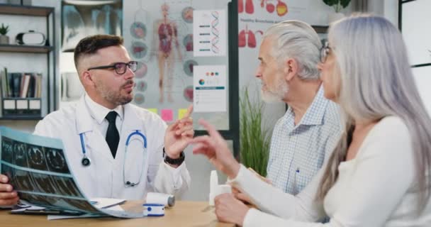 Atractivo médico barbudo positivo que explica los resultados del escáner de rayos X a una pareja mayor segura que lo visita en el consultorio médico — Vídeos de Stock