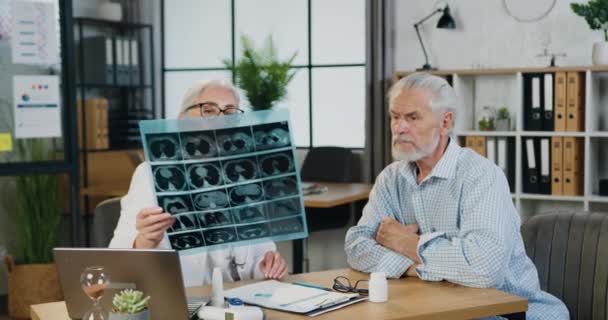 Attraktiv säker respekterad skicklig gråhårig kvinna-läkare förklarar resultaten av röntgen scan för allvarliga mogna skäggiga patienten i moderna medicinska kontor, front view — Stockvideo