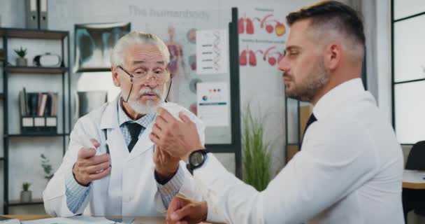 Beau homme barbu concentré prenant des bouteilles avec des pilules d'expérimenté responsable barbu médecin principal et écouter des conseils comment les prendre, vue de face — Video