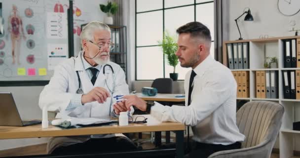 Достойный доверия бородатый 40-летний бизнесмен, посещающий медицинский кабинет, где уважаемый профессиональный зрелый врач измеряет его уровень кислорода специальным датчиком — стоковое видео