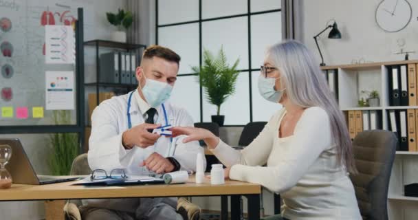 Erfahrener männlicher Arzt in Maske misst den Sauerstoffgehalt älterer Patientinnen im Blut mittels Pulsoximeter während des Krankenhausbesuchs. — Stockvideo