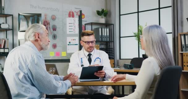 Le médecin traitant en lunettes discute avec un couple marié âgé de l'état de santé lors de l'accueil des patients à l'hôpital — Video