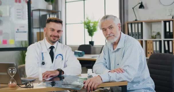 Atraktivní usměvavý vousatý lékař a jeho starší dospělý pacient ukazující palce do kamery během návštěvy kliniky. — Stock video