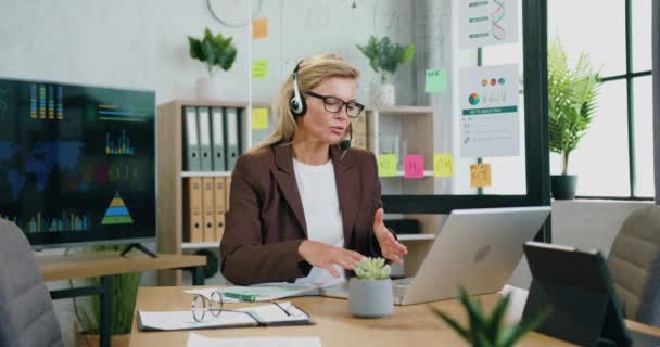 Buena apariencia confiada responsable rubia calificada empresaria en auriculares celebración de una conferencia en línea en el ordenador portátil con los socios simultáneamente tomar notas en el informe — Vídeos de Stock