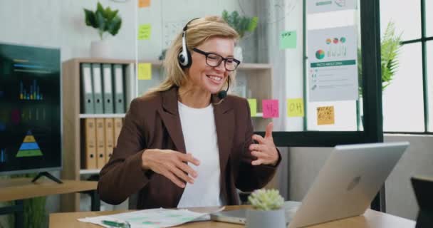 在办公室工作的时候，戴着无线耳机的有经验的金发女商人会通过视频聊天进行愉快的交谈，她笑得很开心 — 图库视频影像