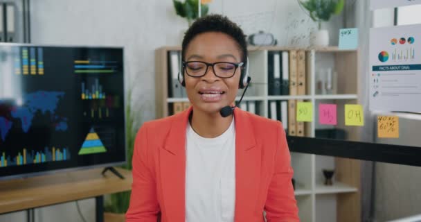 Камера съемки на красивых положительных успешных высококвалифицированных африканских деловых женщин в наушниках, которые разговаривают в камеру во время видеочата с аудиторией — стоковое видео