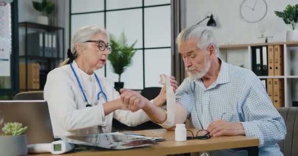 Female doctor removes the bandage from a sprained wrist of an elder male in a hospital room or emergency room — Stock Video