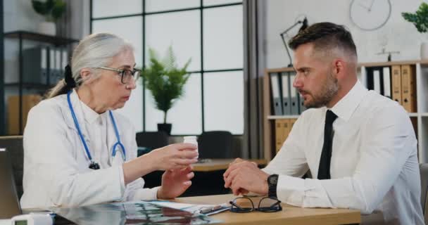 診療所訪問中にデスクでオフィスの薬のボトルについて患者に説明する女性医師 — ストック動画