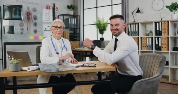 Schöne männliche Patient und ziemlich reife Ärztin suchen Kamera glücklich zeigt Daumen nach oben während hat Besuch in der Klinik — Stockvideo