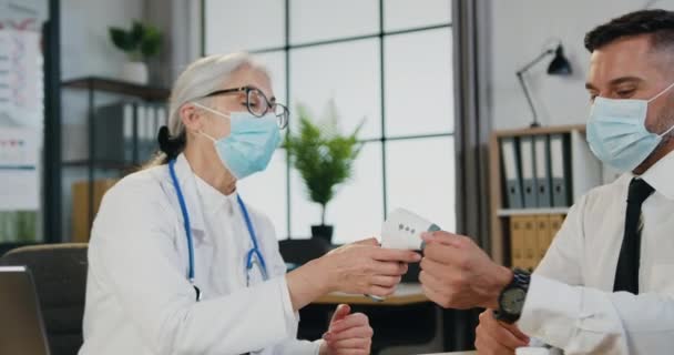 Volwassen vrouwelijke arts in masker is met stethoscoop meten temperatuur haar mannelijke patiënt met infrarood thermometer tijdens afspraak in de medische kliniek — Stockvideo