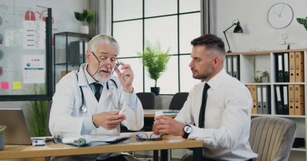 Starší ošetřující lékař vysvětluje předepisování léků pacientovi, zatímco drží lahvičku pilulek sedících v kanceláři u stolu během poradenství na klinice nebo v nemocnici — Stock video