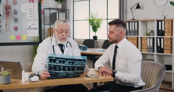 Wykwalifikowany lekarz z brodą w okularach wyjaśniający wyniki badań rentgenowskich pacjenta podczas wizyty w klinice lub szpitalu. Koncepcja opieki zdrowotnej — Wideo stockowe