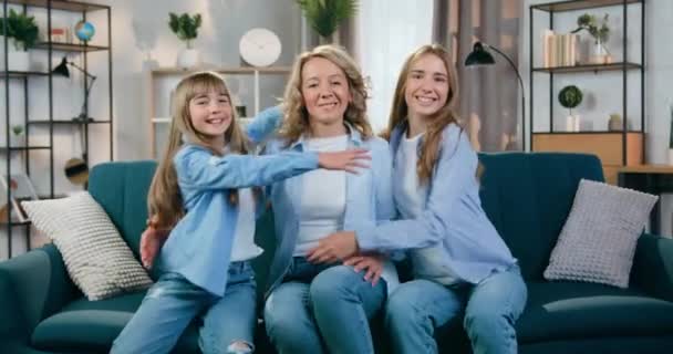 Home portrait of happy adorable smiling mother and her two daughters which sitting on the soft couch in beautifully decorated sitting room and hugging each other — Stock Video