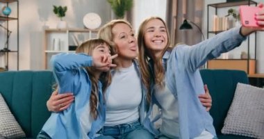 Aile eğlencesi konsepti. Sevimli, mutlu, güler yüzlü sarışın anne ve kızları evde akıllı telefondan komik bir selfie çekerken birlikte eğleniyorlar.