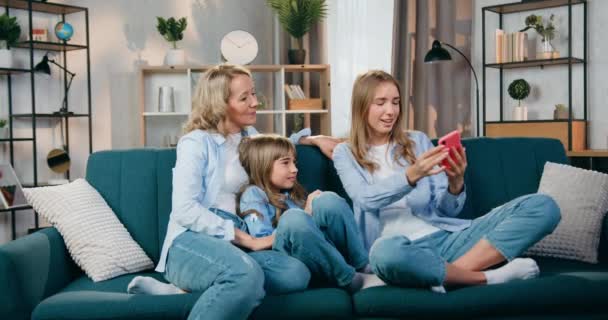 Atraente feliz positiva mãe amigável e suas duas filhas passar o lazer em casa e ter chamada de vídeo no celular, eles acenando as mãos para dizer hallo para parentes — Vídeo de Stock