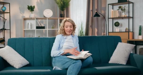 Közelkép elég pozitív nyugodt felnőtt szőke nő, amely könyvet olvas a kanapén otthon, amikor a két lánya rohan, és ad neki ölelést — Stock videók