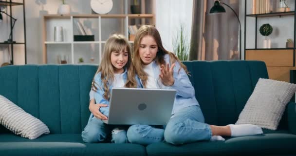 Casa conceito de lazer onde bonito feliz sorrindo despreocupado amigável duas irmãs loiras sentadas no sofá macio em casa durante a chamada de vídeo positivo no laptop — Vídeo de Stock